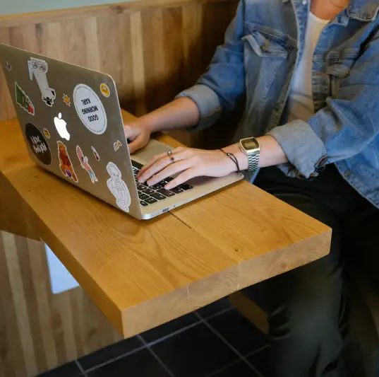 Manos escribiendo en una laptop llena de stickers sobre una mesa de madera.